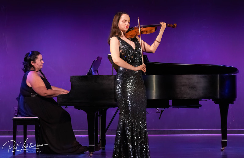 Violinist in a black dress accompanied by a piano player on a stage with a purple background,